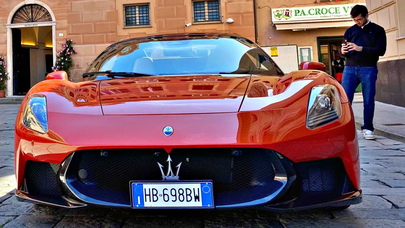 A red Maserati sports car, showcasing MCPura key design elements, is parked on a cobblestone street, with a man nearby using his phone and a building in the background. Q&A: Maserati’s Mastermind Reveals These Standout Features.