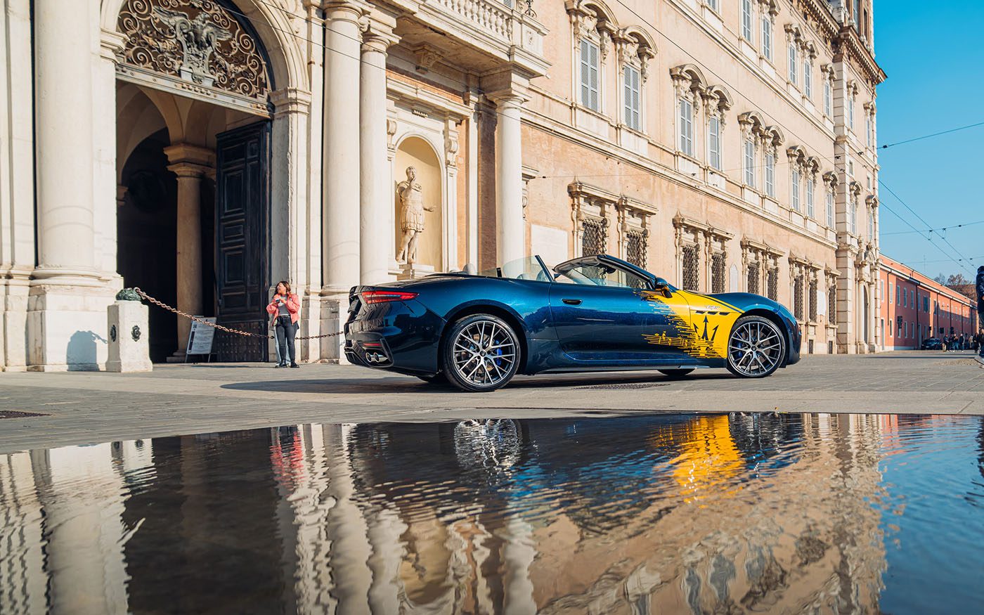 A blue Maserati convertible with yellow detailing is parked outside a grand historic building in Modena, its sleek form elegantly reflected in a puddle on the street.