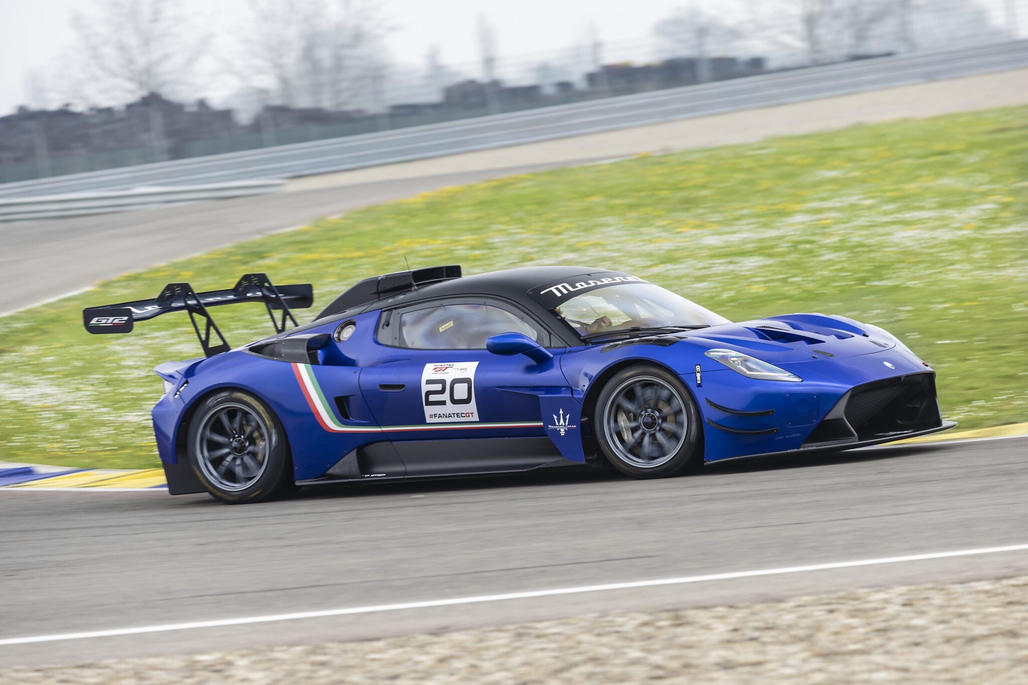 A blue Maserati race car with number 20 on the door drives on a racetrack, equipped with a rear wing and racing decals, competing in the 2026 Championship.