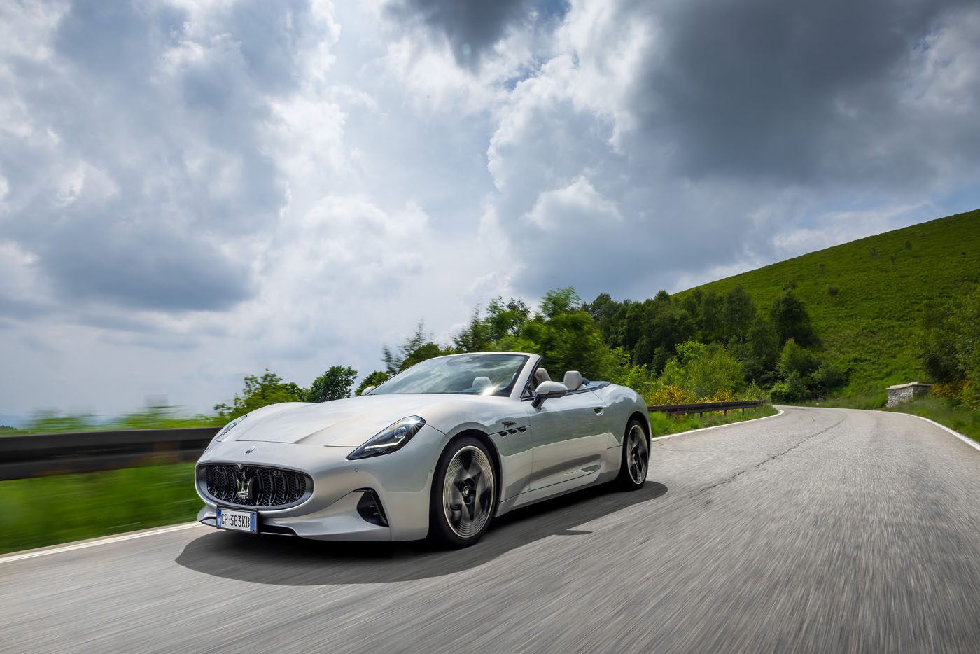 A white convertible sports car drives on a winding road through a green, hilly landscape under a cloudy sky.