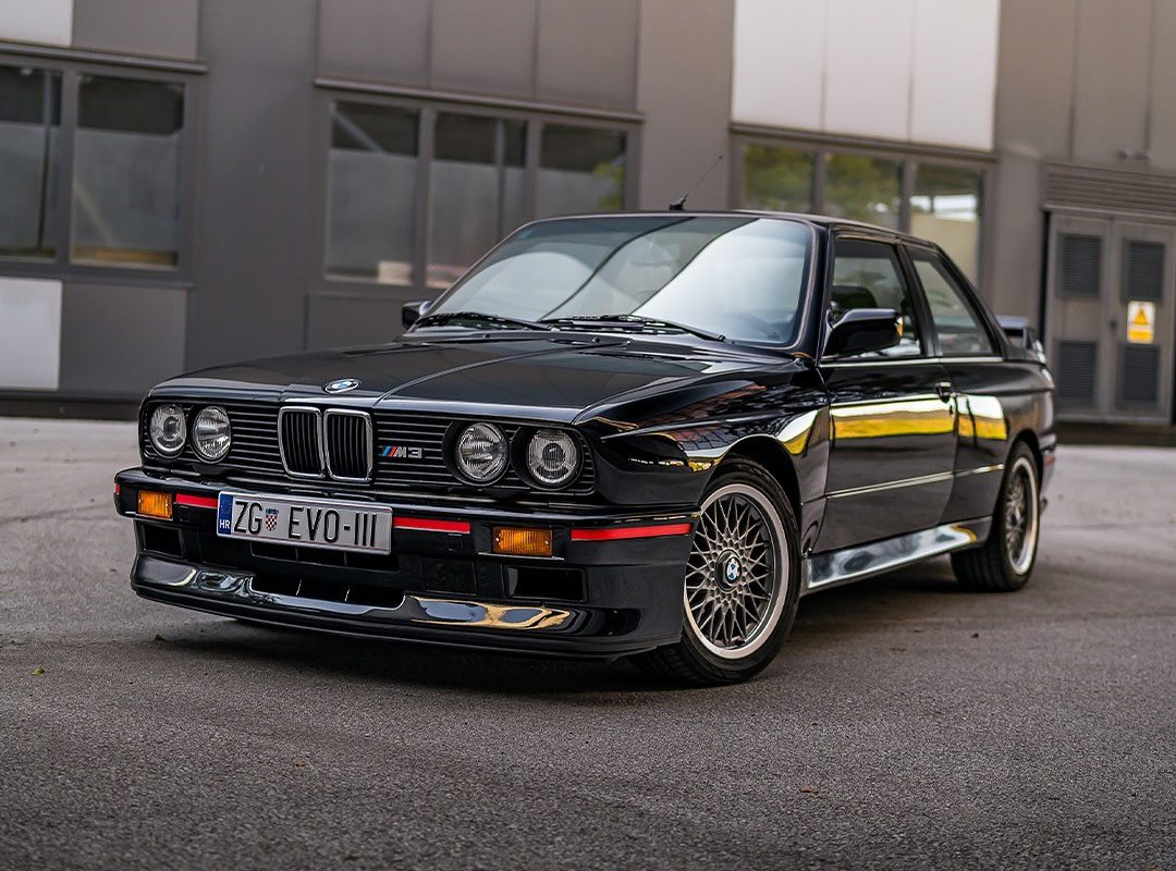 A black BMW E30 M3, a true gem for any car collection, is parked on a paved surface in front of a modern building with reflective windows and a license plate reading "ZG EVO-III.