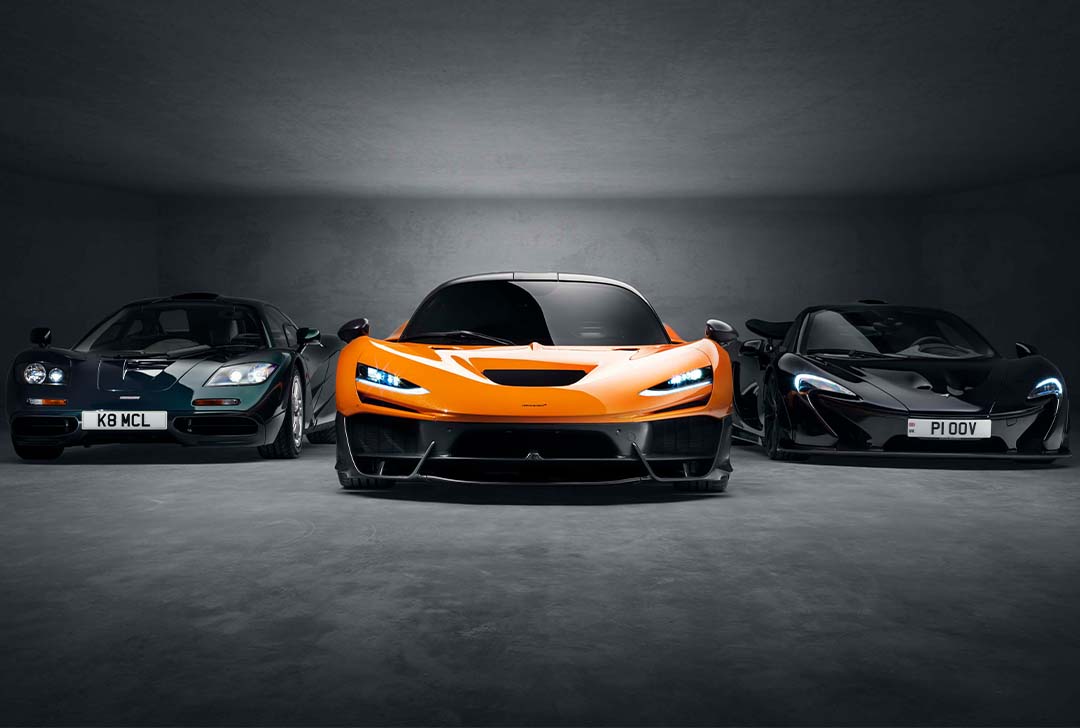 Three McLaren sports cars—a classic F1, a bright orange modern McLaren W1, and a sleek black model—are parked side by side in a dimly lit space reminiscent of Goodwood’s exclusive showcases.