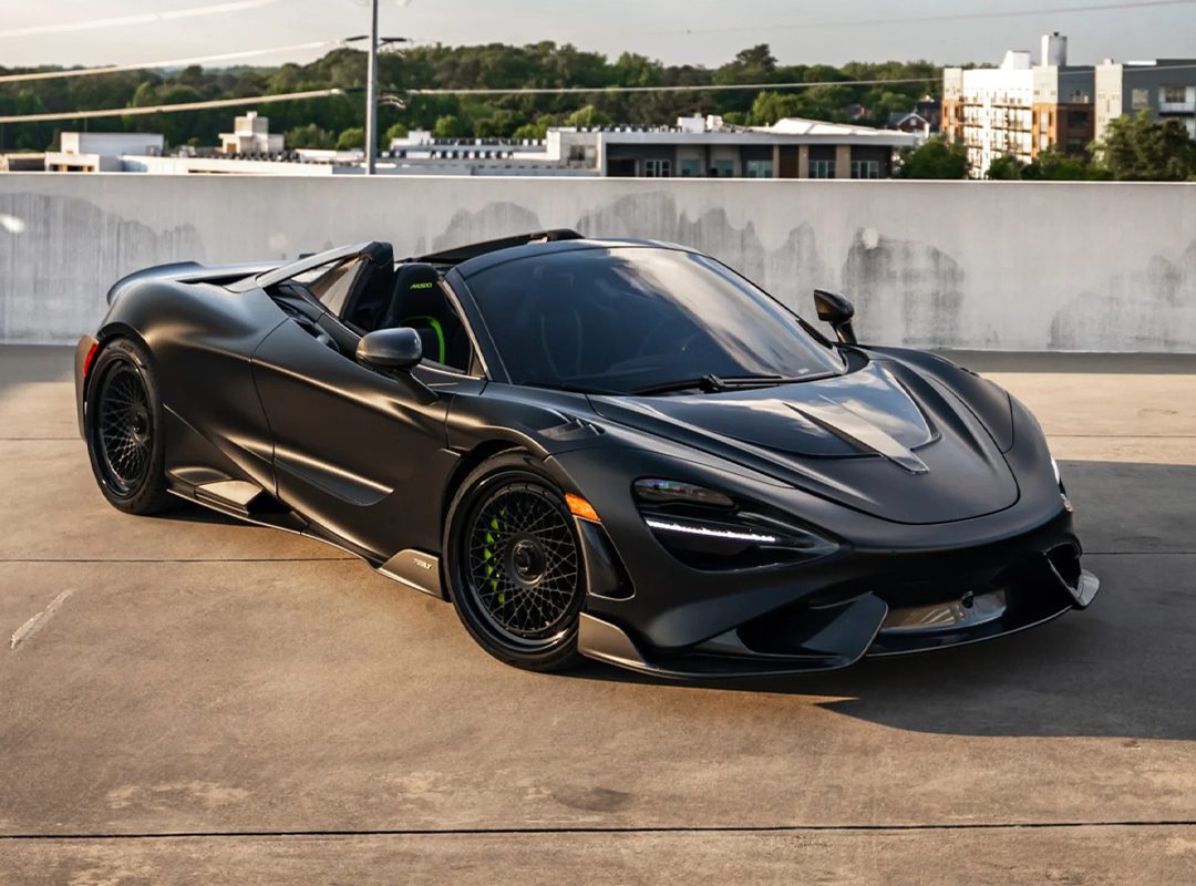 A matte black McLaren 720S Spider convertible in stealth mode is parked on a rooftop, with buildings and trees in the background—perfect for those seeking exclusive matte black cars for sale.
