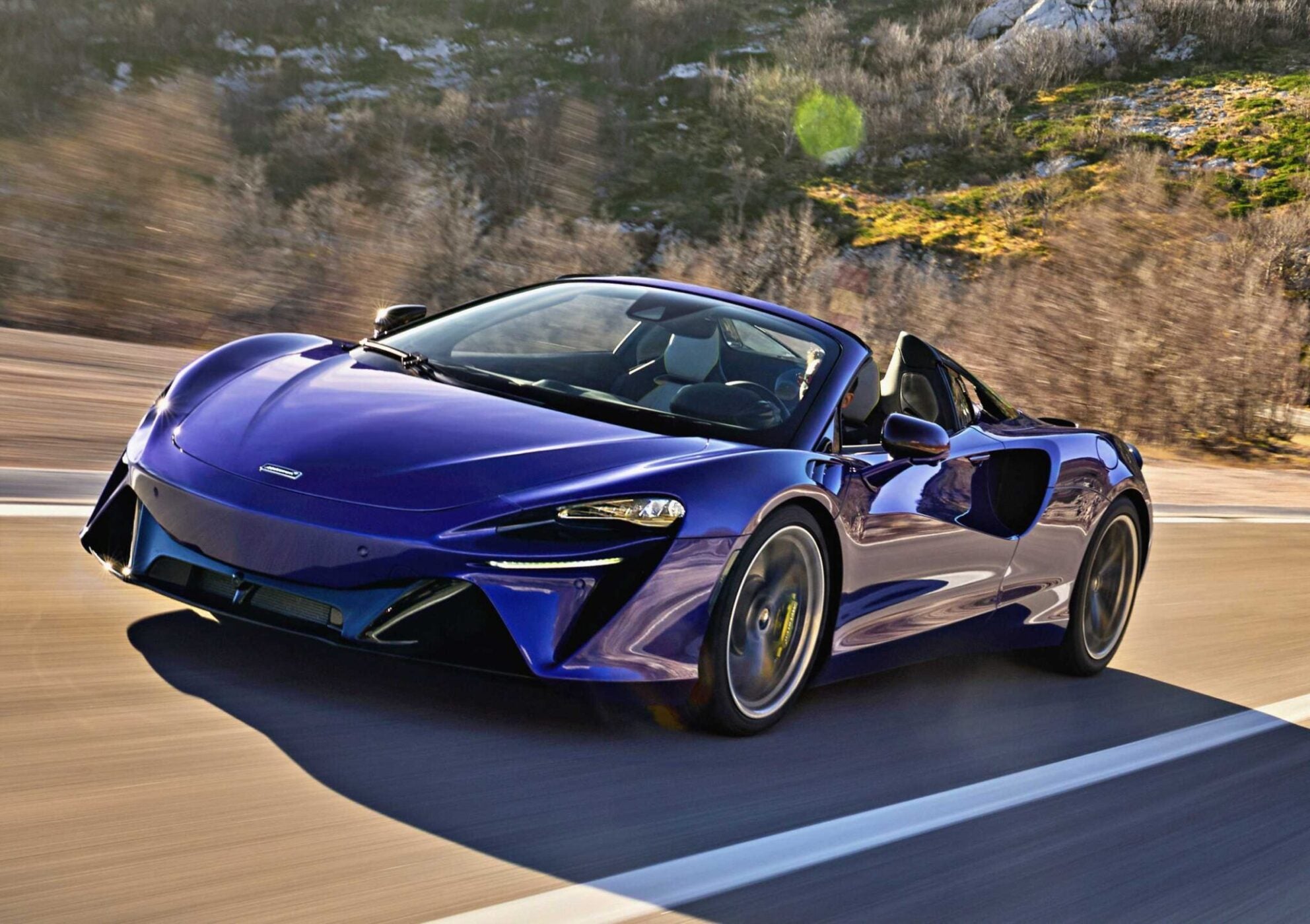 A blue convertible sports car, like the 2025 McLaren Artura Spider, speeds along a road with blurred trees and rocky terrain in the background—perfectly capturing the droptop’s split personality in motion.