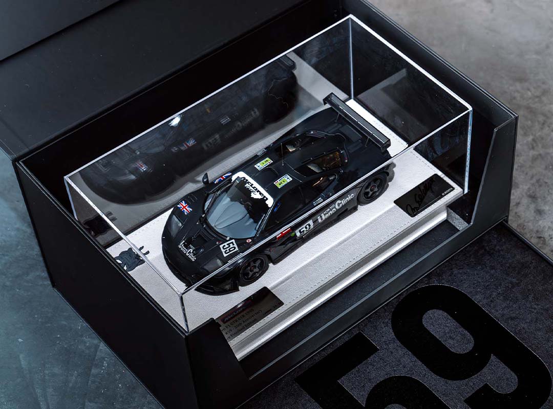 A black McLaren F1 GTR model race car with the number 59 is displayed in a glass case with a white base, inside a black Lanzante box featuring the number 59, celebrating its iconic Goodwood legacy.