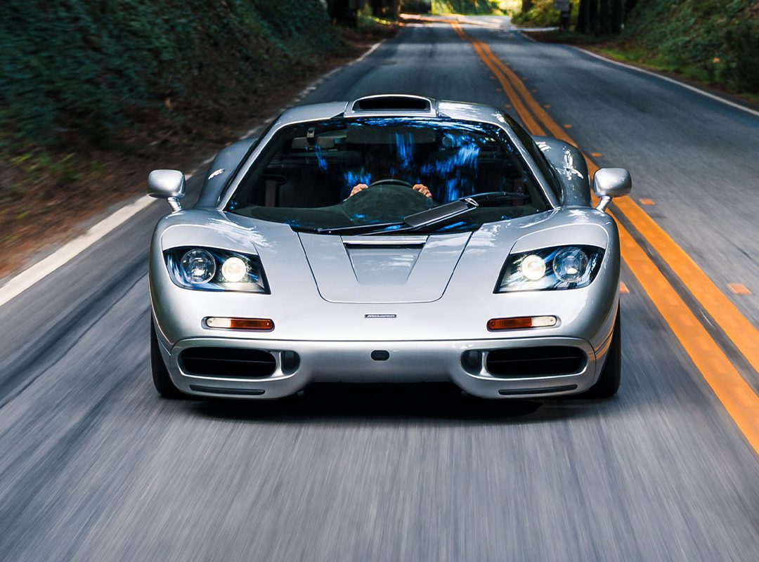A silver McLaren F1 sports car drives down a two-lane road surrounded by greenery, viewed from the front—an iconic sight reminiscent of RM Sotheby's Monterey Auction showcases.