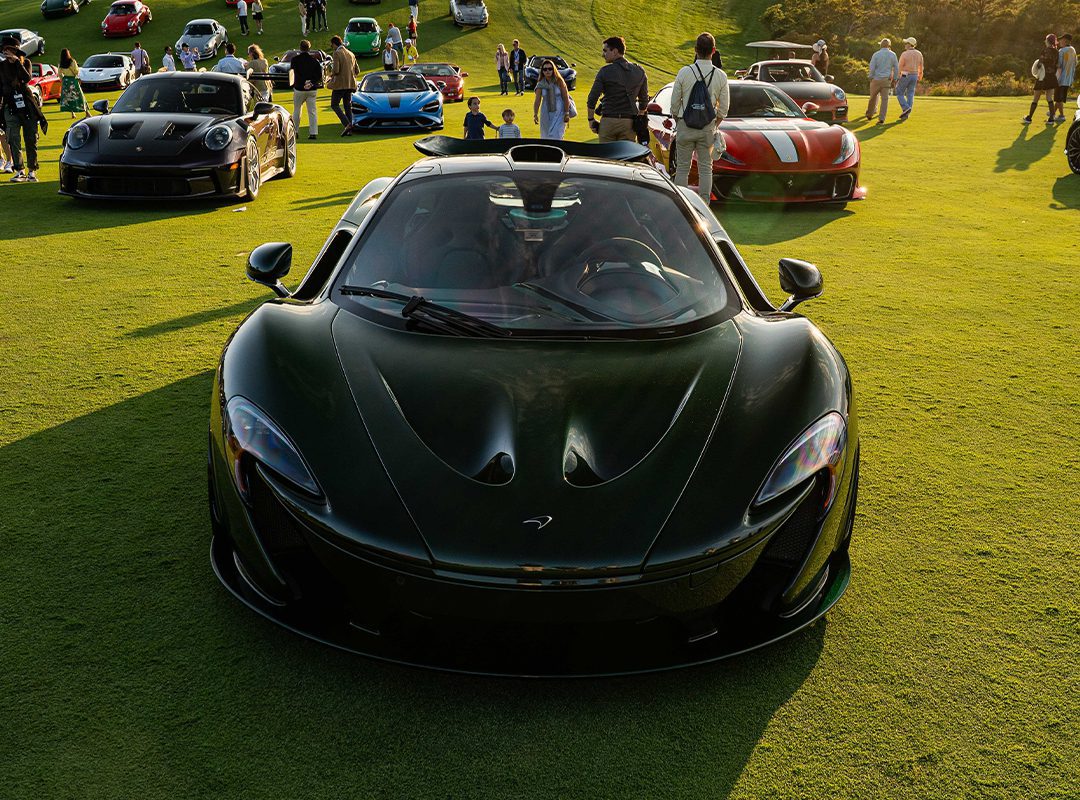 A black McLaren sports car, a true piece of automotive art, is displayed on a grassy field among other luxury vehicles and guests at the exclusive Bridge IX outdoor car event.
