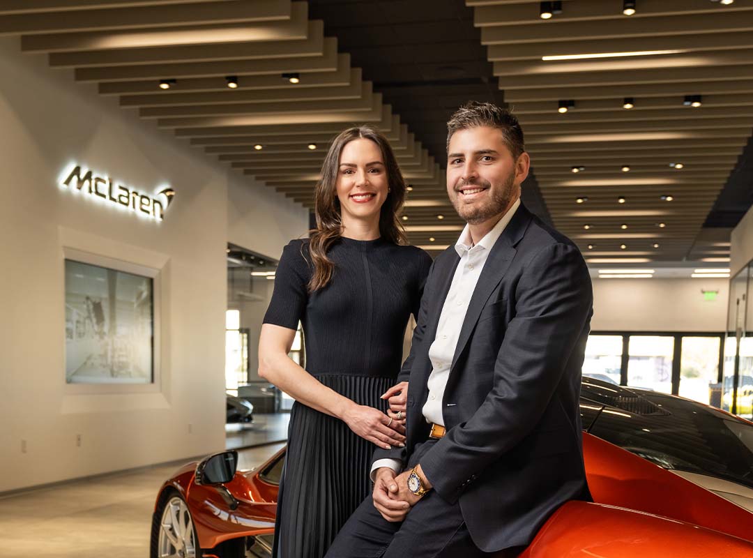 A man and woman pose together inside McLaren Scottsdale, standing in front of an orange sports car, capturing a memorable moment in this Year of Growth.
