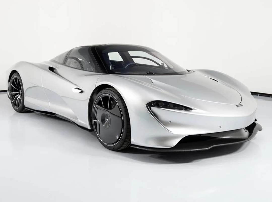 A silver McLaren Speedtail hypercar, inspired by the Electrified Longtail concept, is parked in a white studio, featuring a sleek, aerodynamic design with black accents and futuristic wheels.