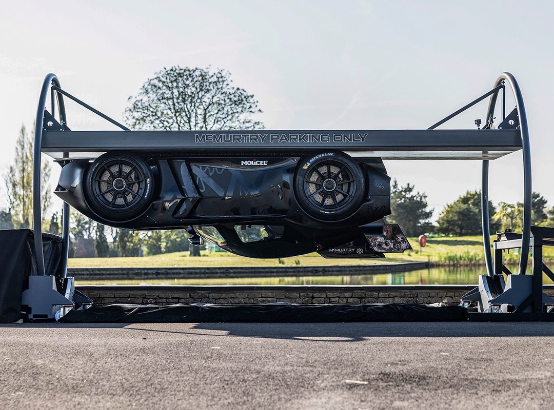 A McMurtry Spéirling is parked horizontally on a raised platform labeled "McMurtry Parking Only," creating the illusion of an almost gravity-defying stunt against a serene backdrop of trees and a pond.
