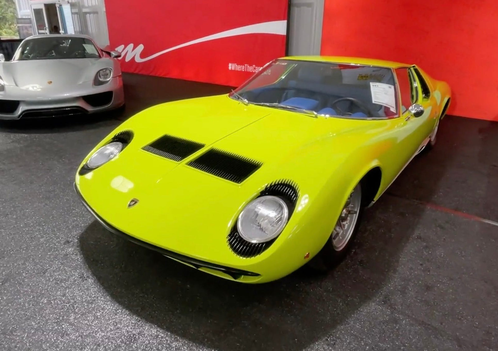 A bright yellow Lamborghini Miura is displayed indoors next to a silver sports car, with a red wall in the background—an iconic pairing reminiscent of the standout vehicles at the 2025 Monterey Mecum Auction: Top 10 Results And Highlights.