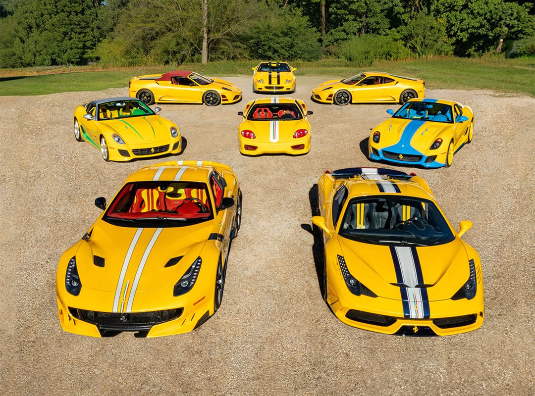Eight yellow Ferrari sports cars with white and blue racing stripes, part of exclusive Ferrari Collections, are arranged in a semicircle on a gravel surface, with trees and grass in the background.