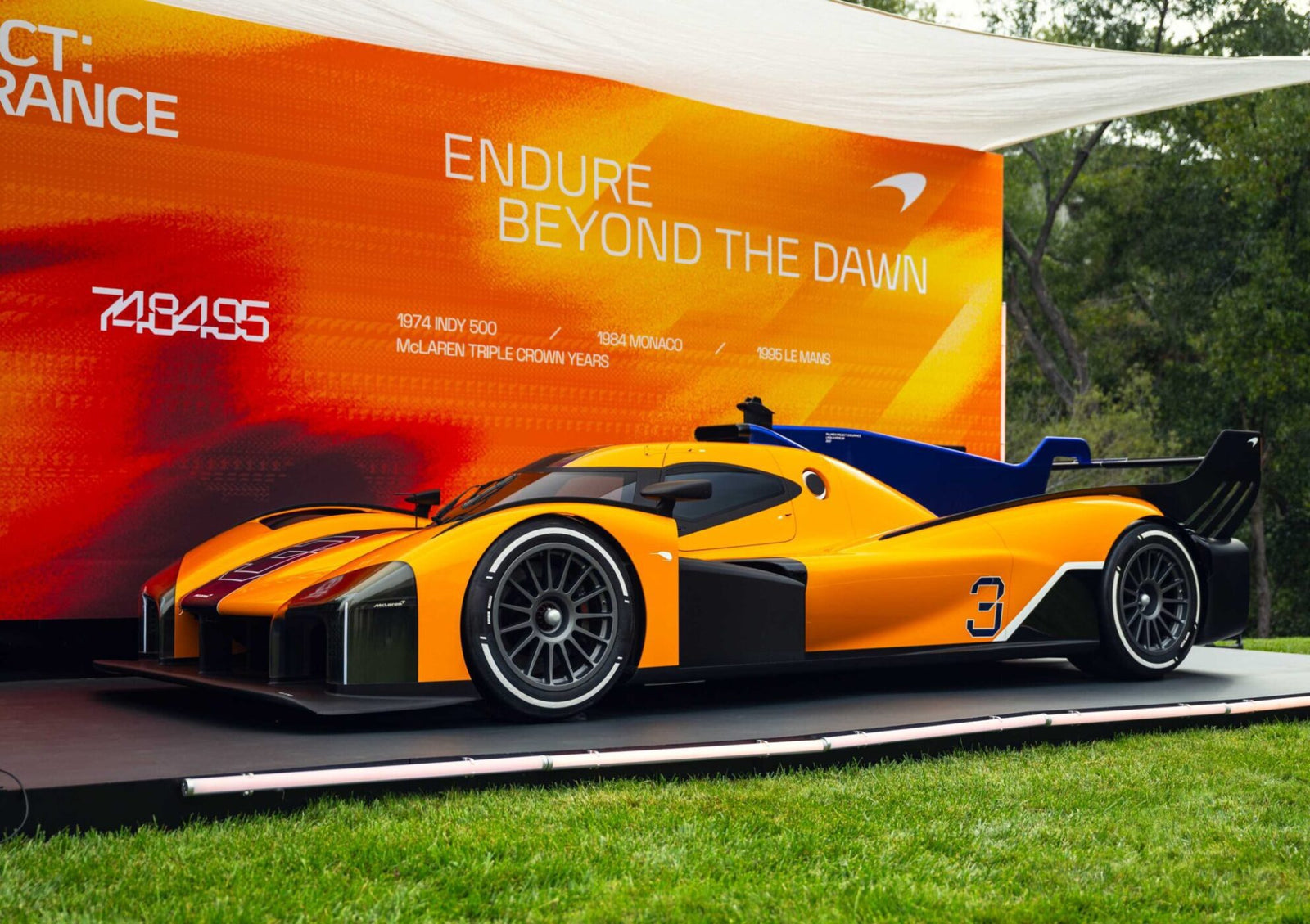 An orange and blue race car is displayed on a platform in front of a vibrant orange McLaren backdrop that reads “ENDURE BEYOND THE DAWN,” celebrating McLaren’s Project: Endurance Hypercar Debuts Triple Crown Livery at Monterey.