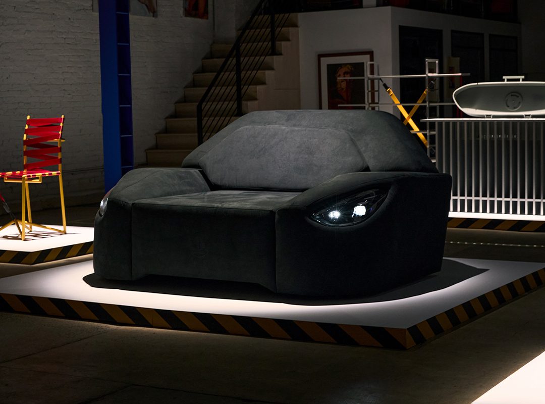 A black sofa designed to resemble the front of a car, complete with headlight-shaped lights and inspired by Mercedes-AMG performance car parts, is displayed on a platform in an indoor setting.