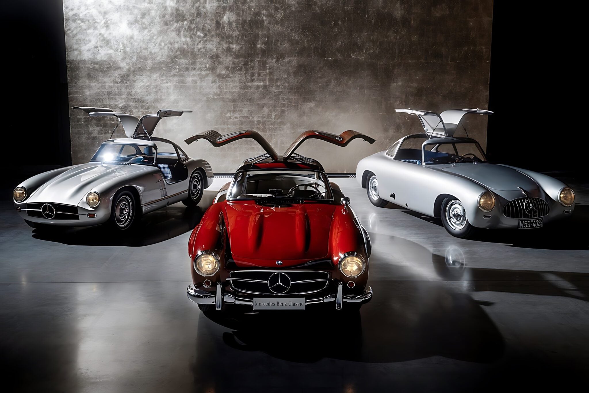 Three classic Mercedes-Benz cars with gullwing doors open, including the iconic 300 SL—the Blue Chip Benz—are displayed in a dimly lit showroom with a textured wall background.