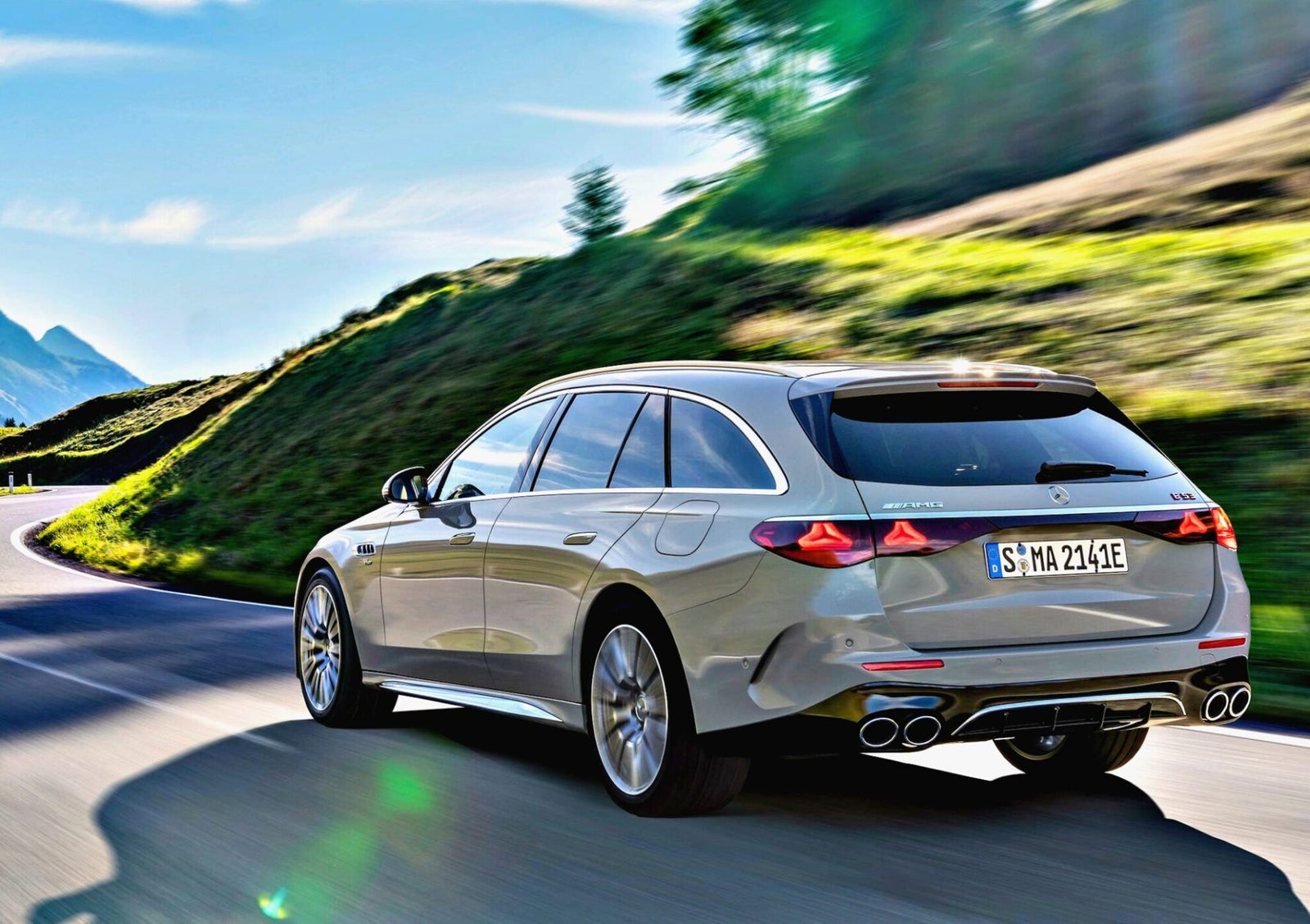 A silver Mercedes-Benz station wagon glides along a winding mountain road under a clear blue sky, with lush green hills in the background, just as Mercedes-AMG reveals pricing for the new E 53 HYBRID Wagon.