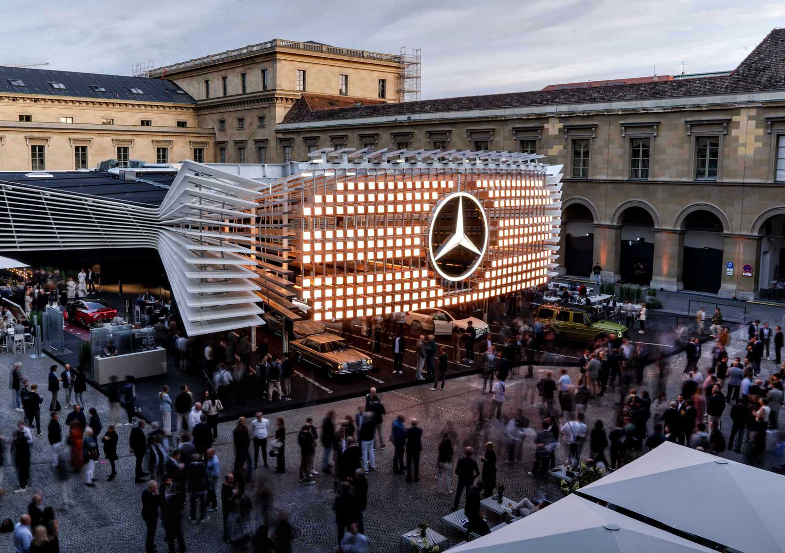 A large crowd gathers around a modern Mercedes-Benz pavilion with an illuminated logo in a historic plaza during an outdoor event, exploring what's Beyond the All-electric GLC: Mercedes-Benz at IAA 2025.