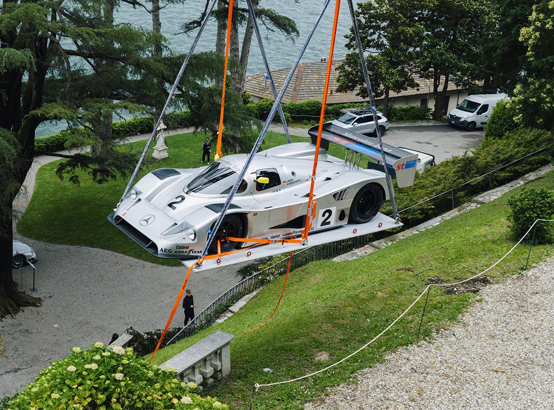 A silver Mercedes-Benz CLK GTR race car is being lifted by orange straps and a crane over a grassy hill, near a driveway with trees and parked vehicles in the background, possibly for FuoriConcorso 2025.
