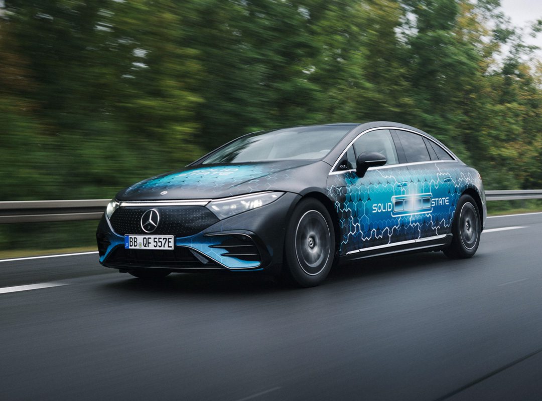 A Mercedes-Benz EQS electric sedan with "Solid State" graphics on the side, highlighting its solid-state battery and impressive 749-mile range, drives on a highway lined with trees in the background.