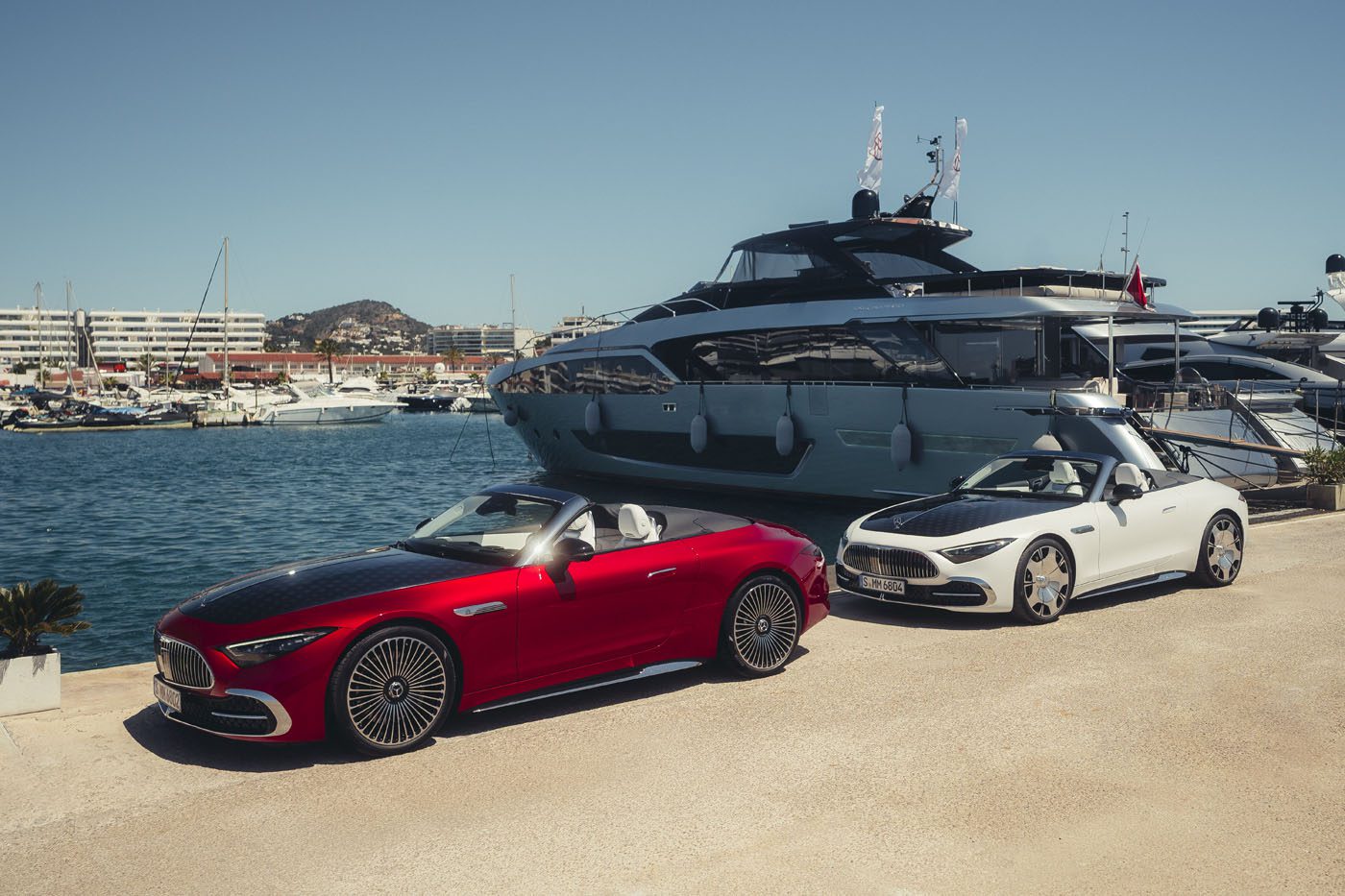 Two modern convertibles, including a striking Mercedes-Maybach SL 680, are parked on a marina dock before a luxury yacht, with other boats and waterfront buildings in the background. The 2026 Mercedes-Maybach will soon be available at select US dealers.
