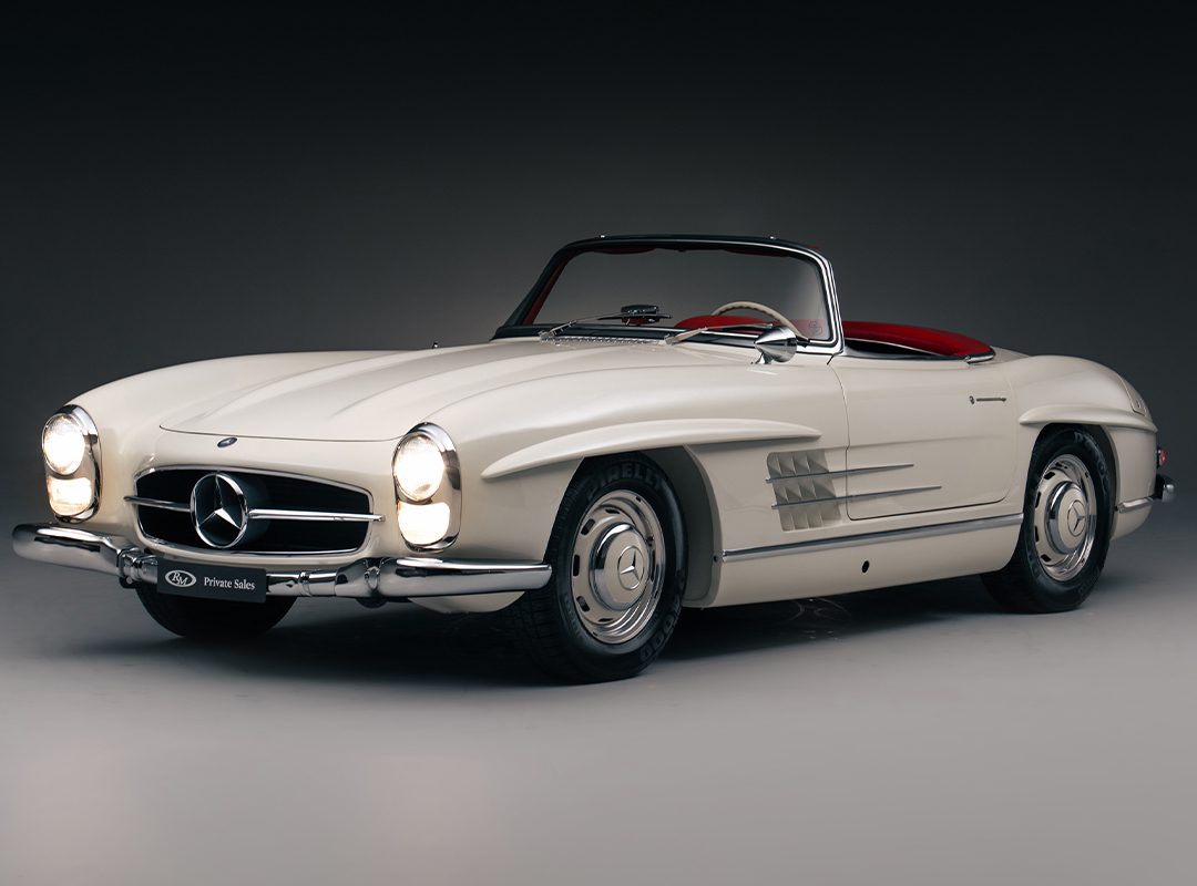A white vintage Mercedes-Benz convertible with red interior, viewed from the front left side against a dark background—a true icon among classic cars.