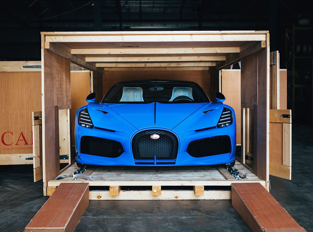 A Bugatti Mistral in striking French Racing Blue sits poised inside a wooden shipping crate, with its sleek front facing outward.