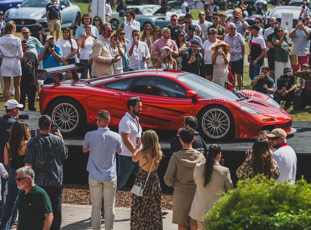 A red sports car is displayed on a platform at duPont REGISTRY's Ultimate Weekend Guide event, with a crowd of people taking photos and admiring the sleek vehicle.
