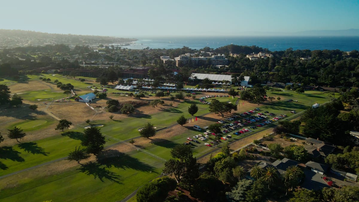 Aerial view of a golf course with scattered trees, parked cars, tents, and nearby buildings; coastline and ocean visible in the background during Mecum Auctions at Monterey Car Week.
