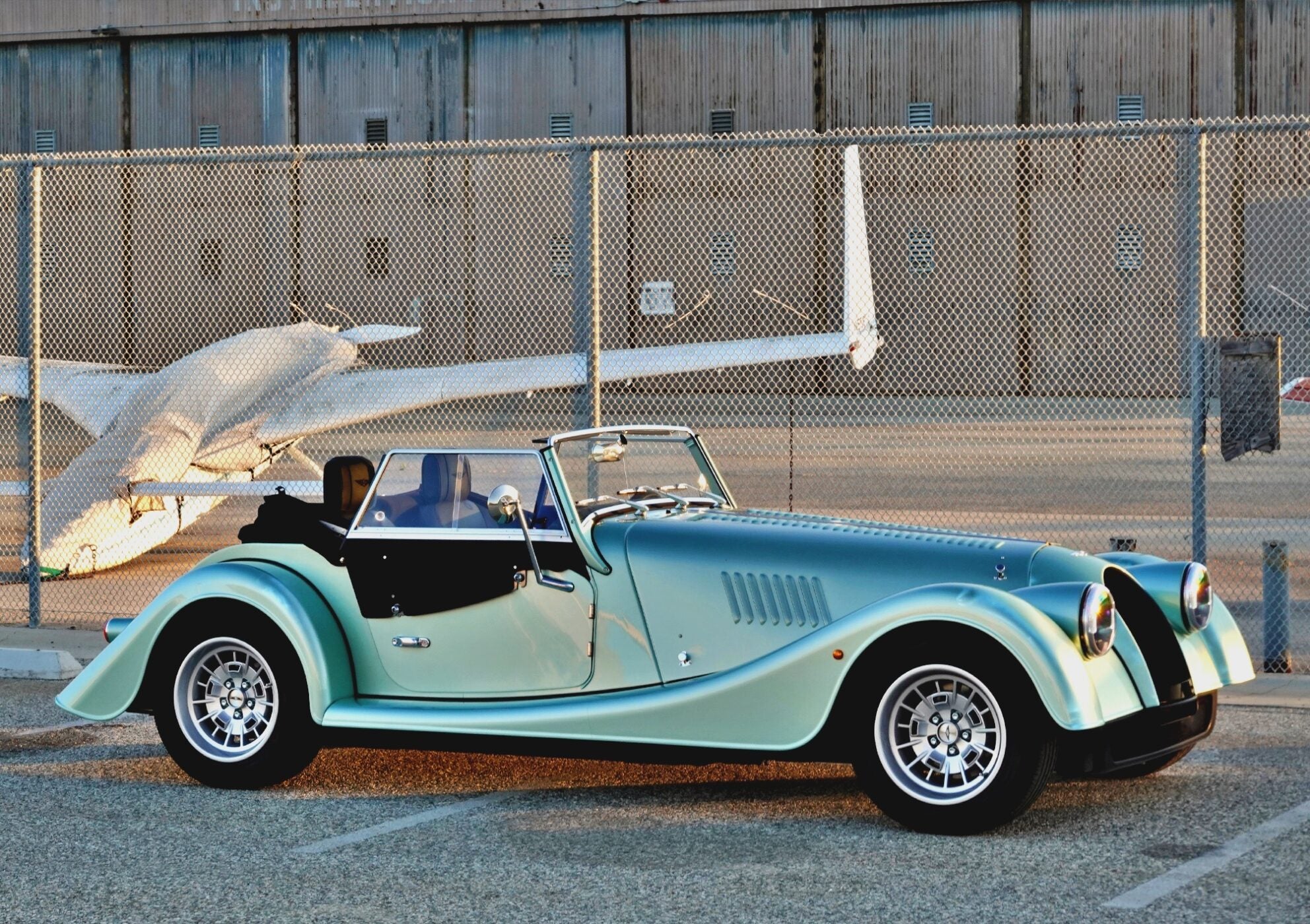 A classic convertible sports car with a teal and black finish, reminiscent of the 2025 Morgan Plus Four Review: A Timeless Reminder of Olde-School Driving Fun, is parked in front of a chain-link fence and a small airplane at an airport.
