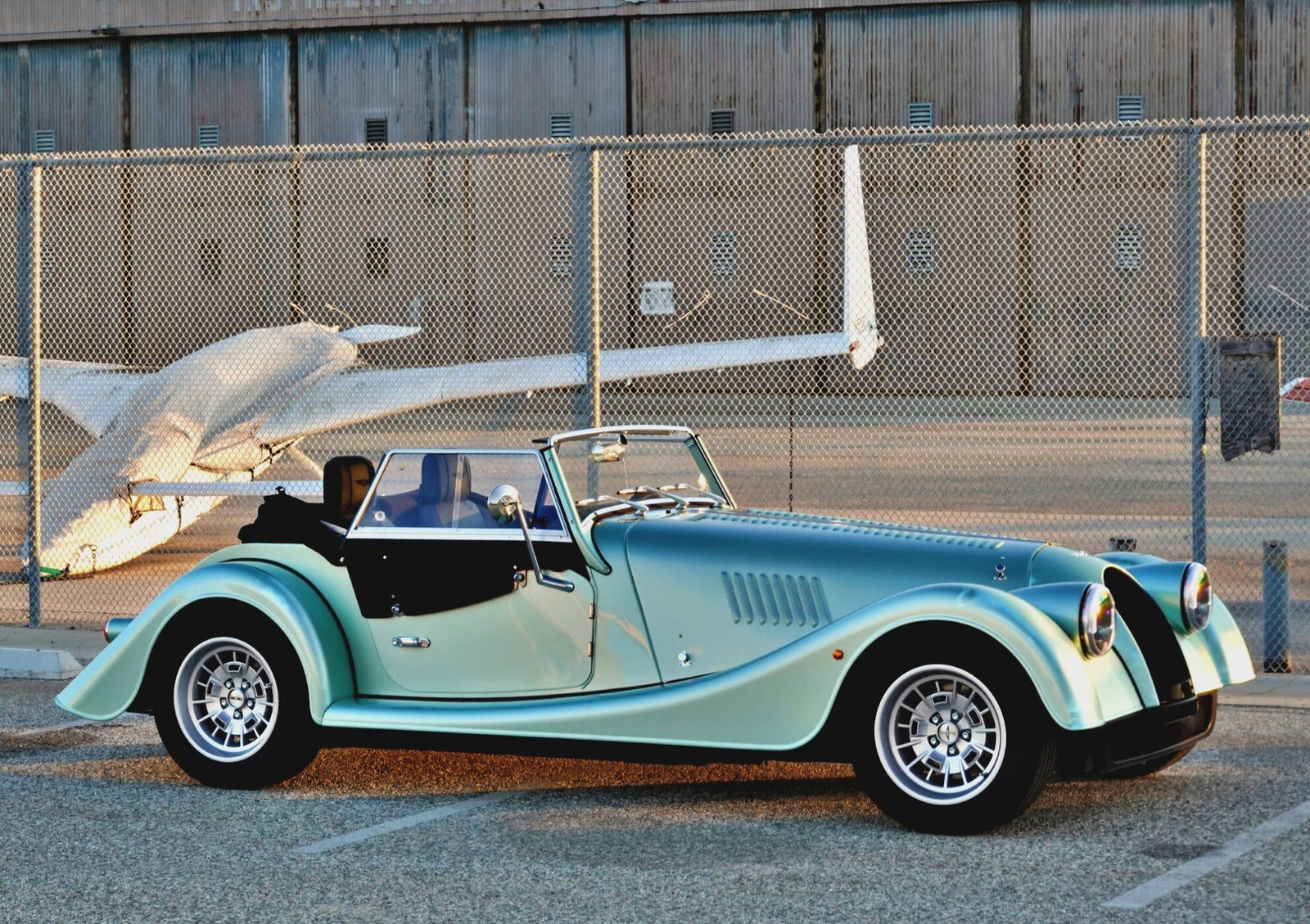 A classic convertible sports car with a teal and black finish, reminiscent of the 2025 Morgan Plus Four Review: A Timeless Reminder of Olde-School Driving Fun, is parked in front of a chain-link fence and a small airplane at an airport.