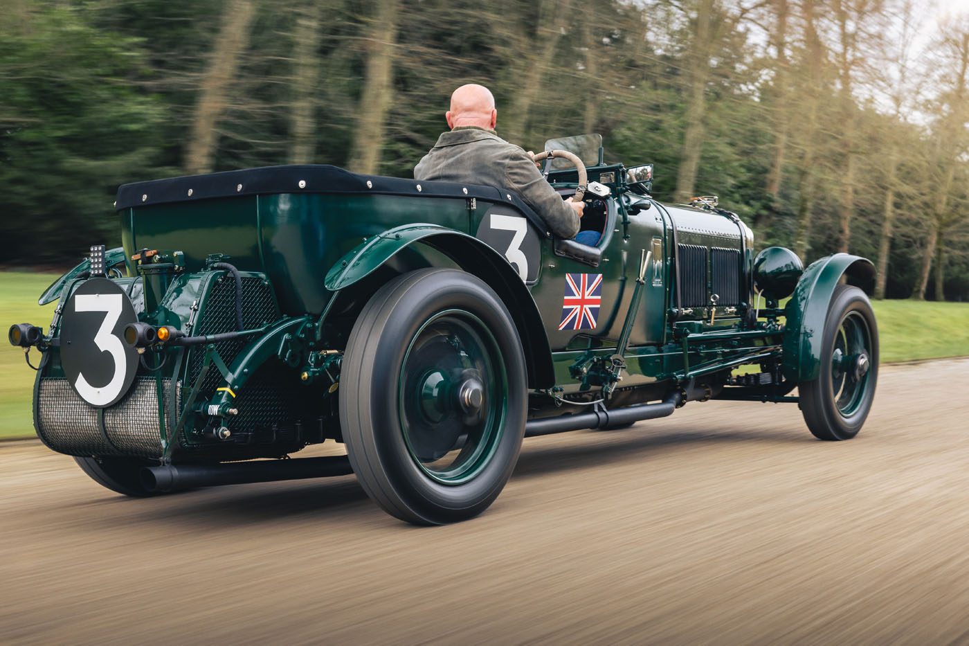 A vintage green Bentley Speed Six from the Continuation Series, with the number 3 and a British flag on the side, is being driven on a road by a man. The classic car features spoked wheels and a rear-mounted spare tire.