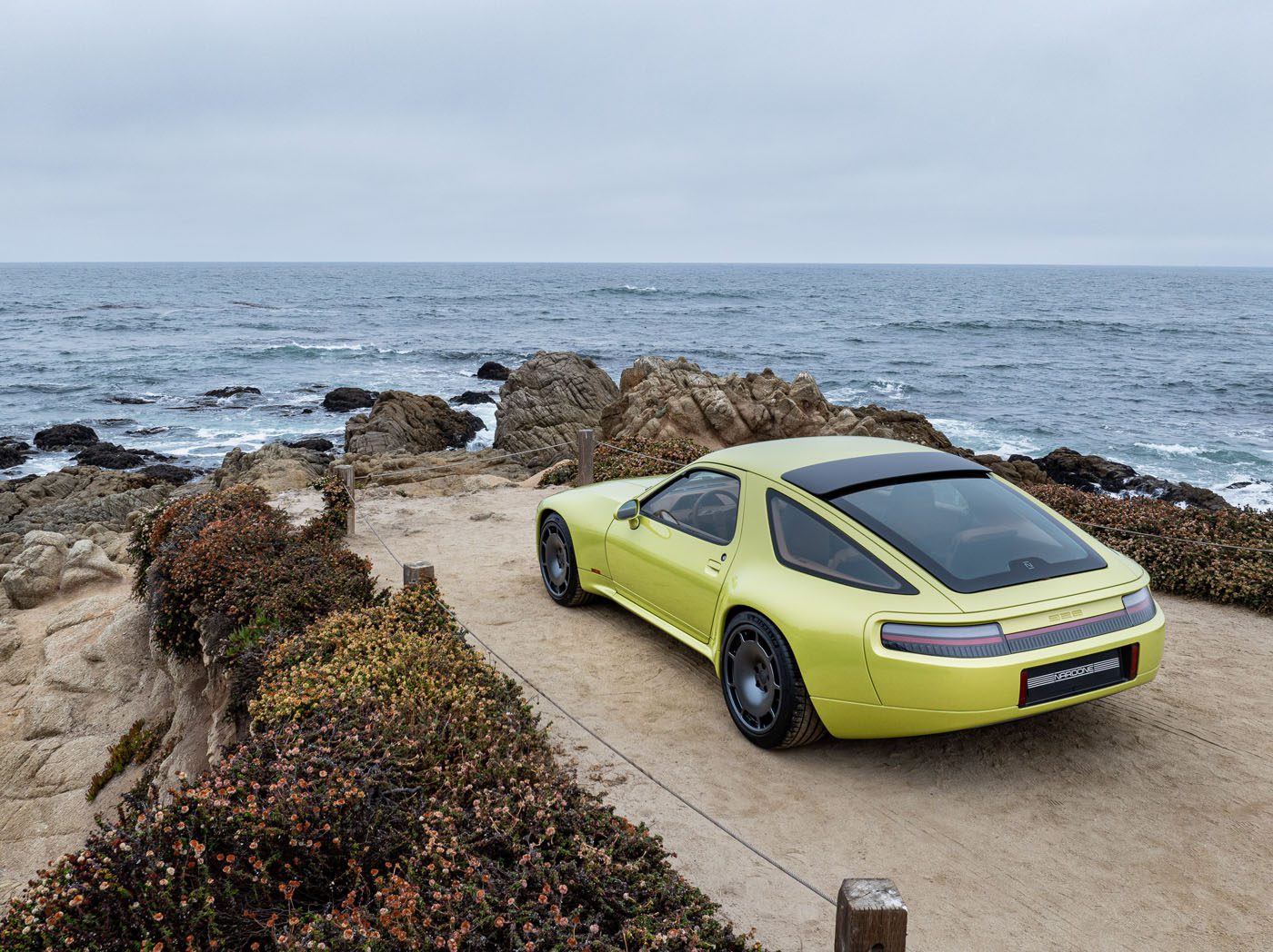 A yellow Nardone 928 sports car is parked on a dirt path overlooking rocky cliffs and the ocean under a cloudy sky.