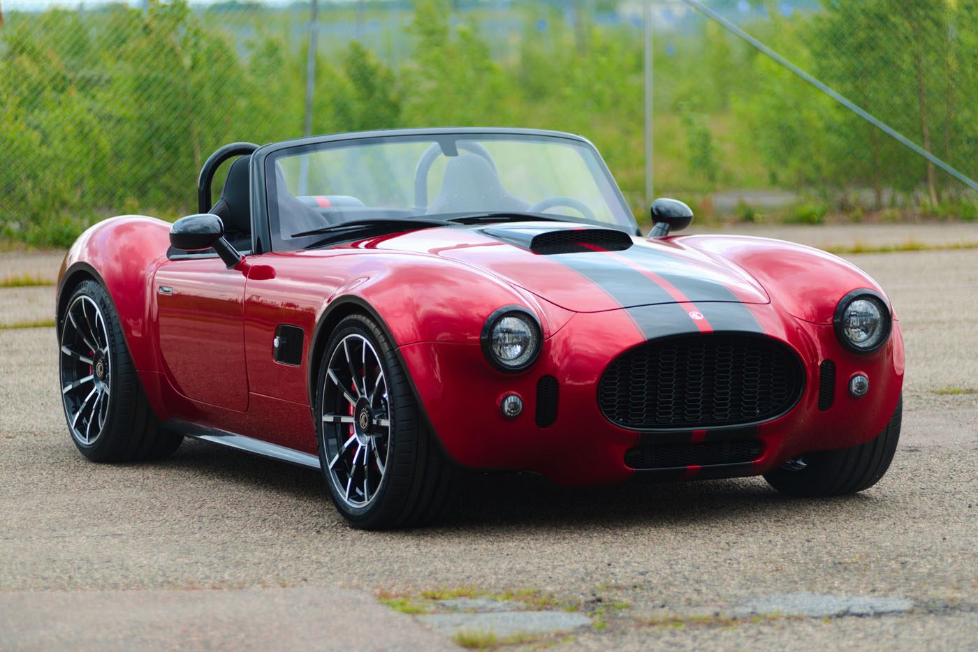 A red Cobra GT Roadster by AC Cars, featuring black racing stripes, is parked on a paved surface, with greenery and a chain-link fence in the background—a true production classic.