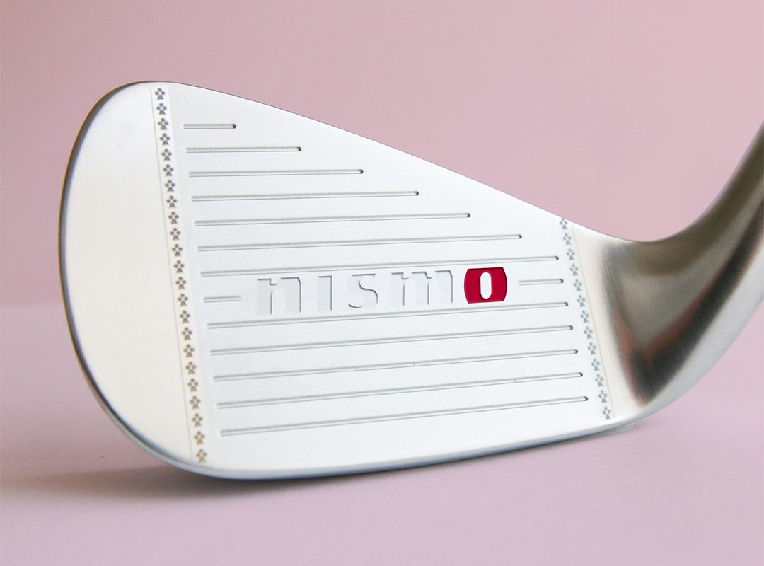 Close-up of a metallic golf club head with the word "nismo" and a red square engraved on the face, set against a pink background.