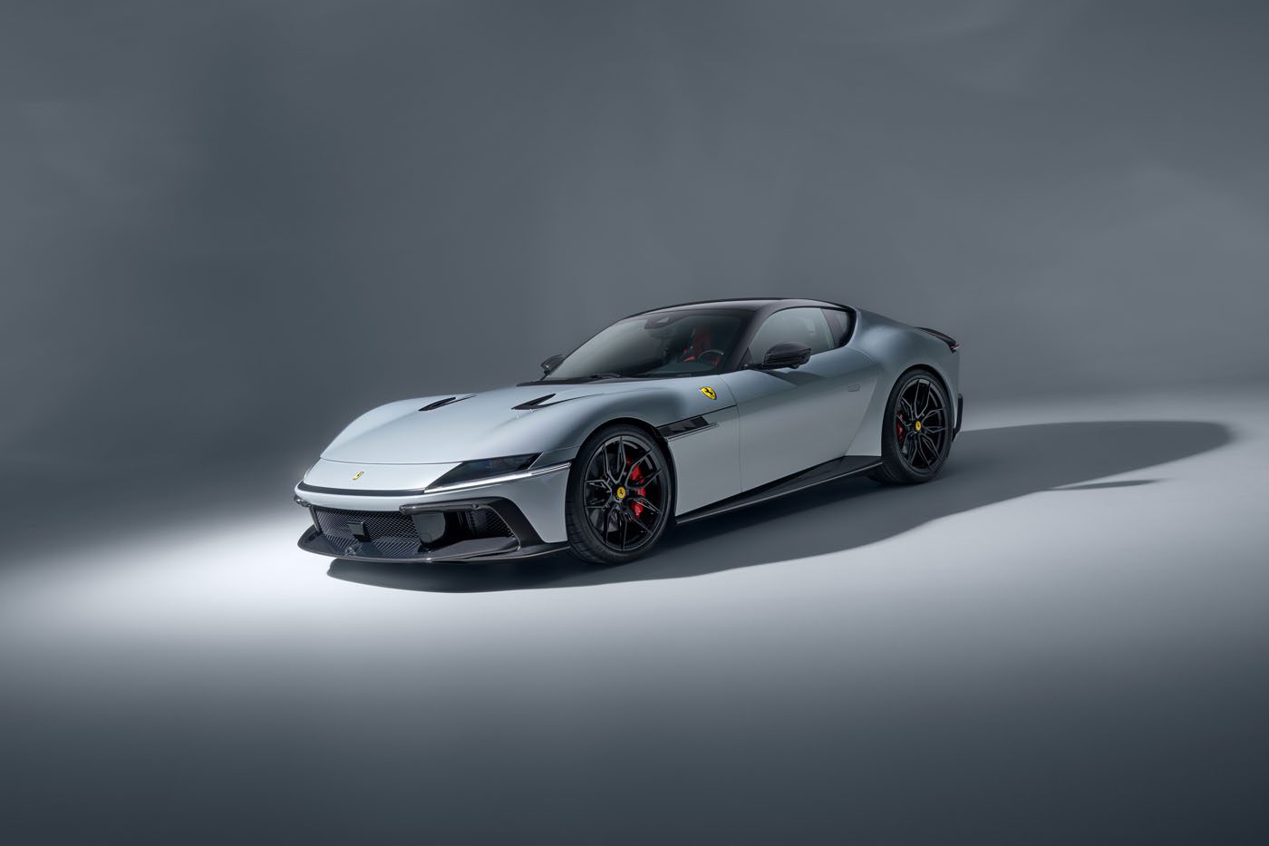 A silver Ferrari sports car is parked on a plain, light-gray studio background, shown from a front three-quarter angle.