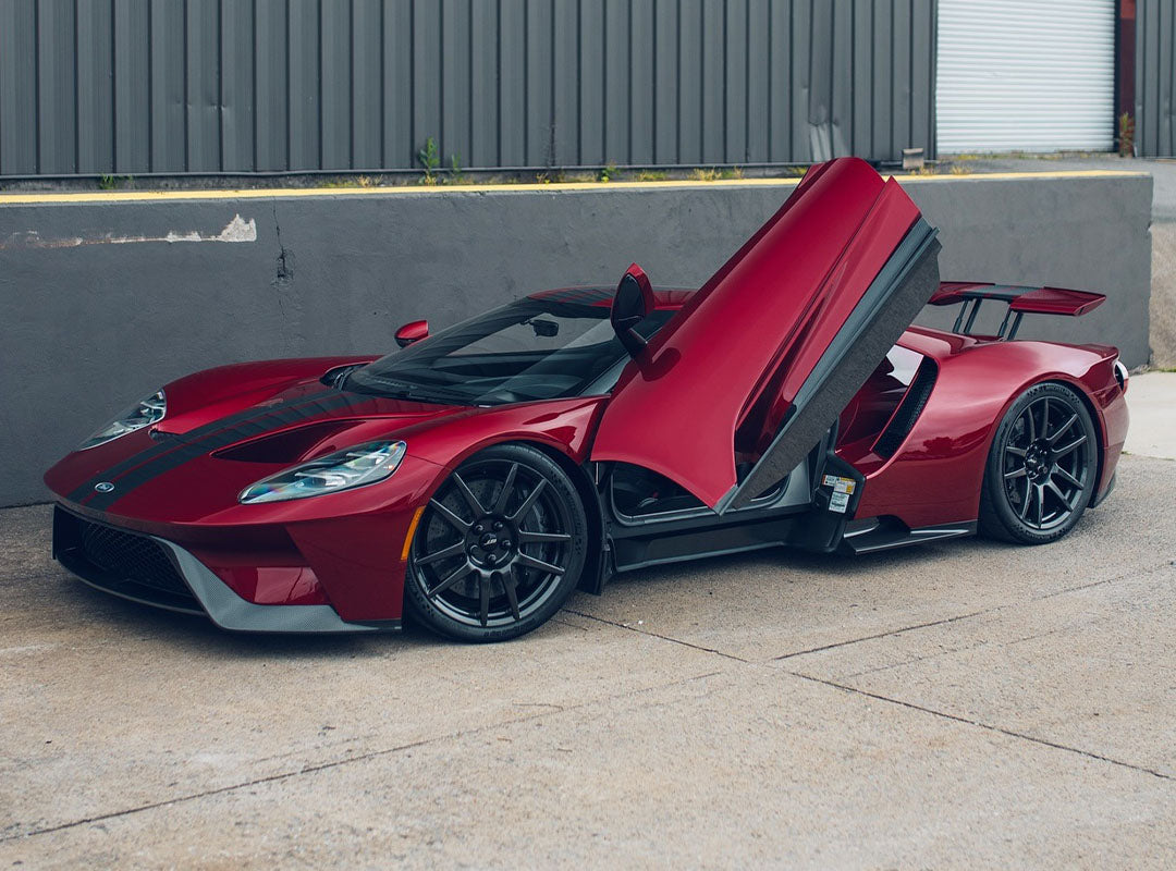 A red exotic car with an upward-opening driver door is parked on a concrete surface beside a gray industrial building, exemplifying the luxury cars offered by Velocity Motorcars.