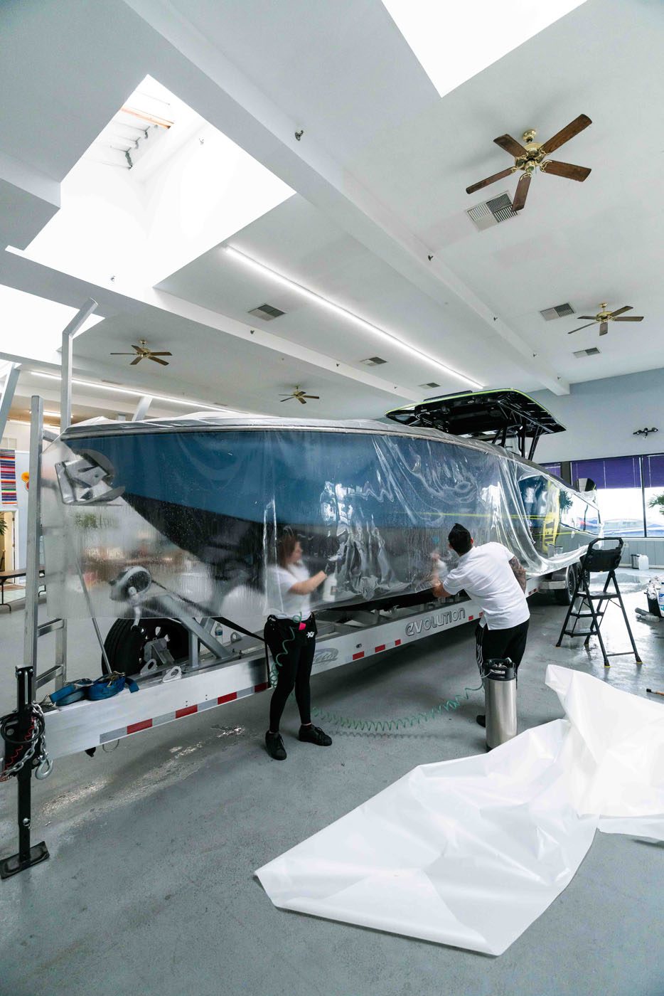 Two people apply Marine Protection Film to a large boat on a trailer inside a spacious, well-lit room with ceiling fans, ensuring optimal vessel protection and marine preservation.