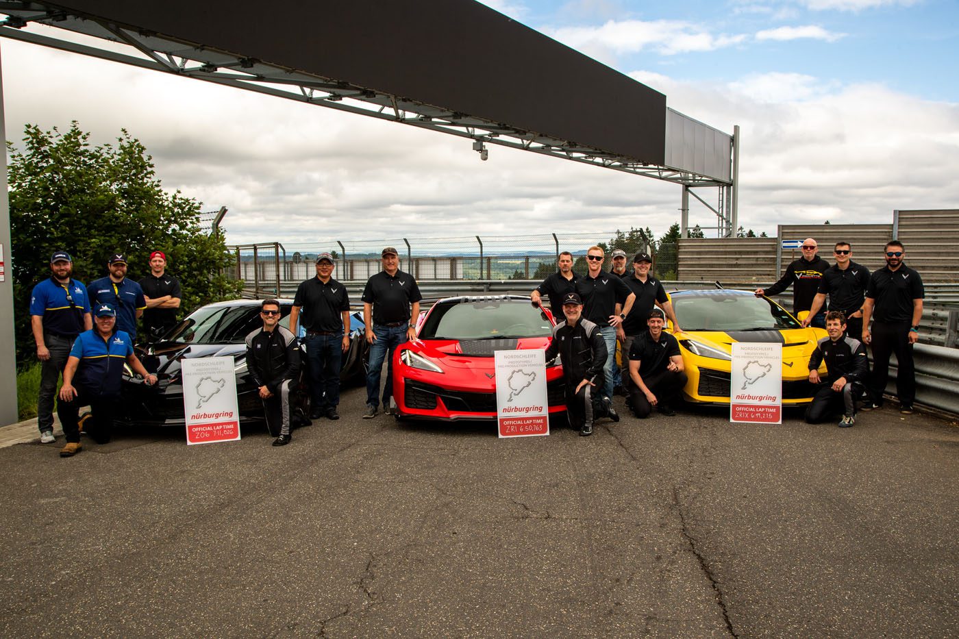 A group of people in black outfits pose beside red, yellow, and blue sports cars—including the Corvette ZR1—on a race track, holding Nürburgring lap time signs touting one of the fastest American production cars’ Nürburgring record.