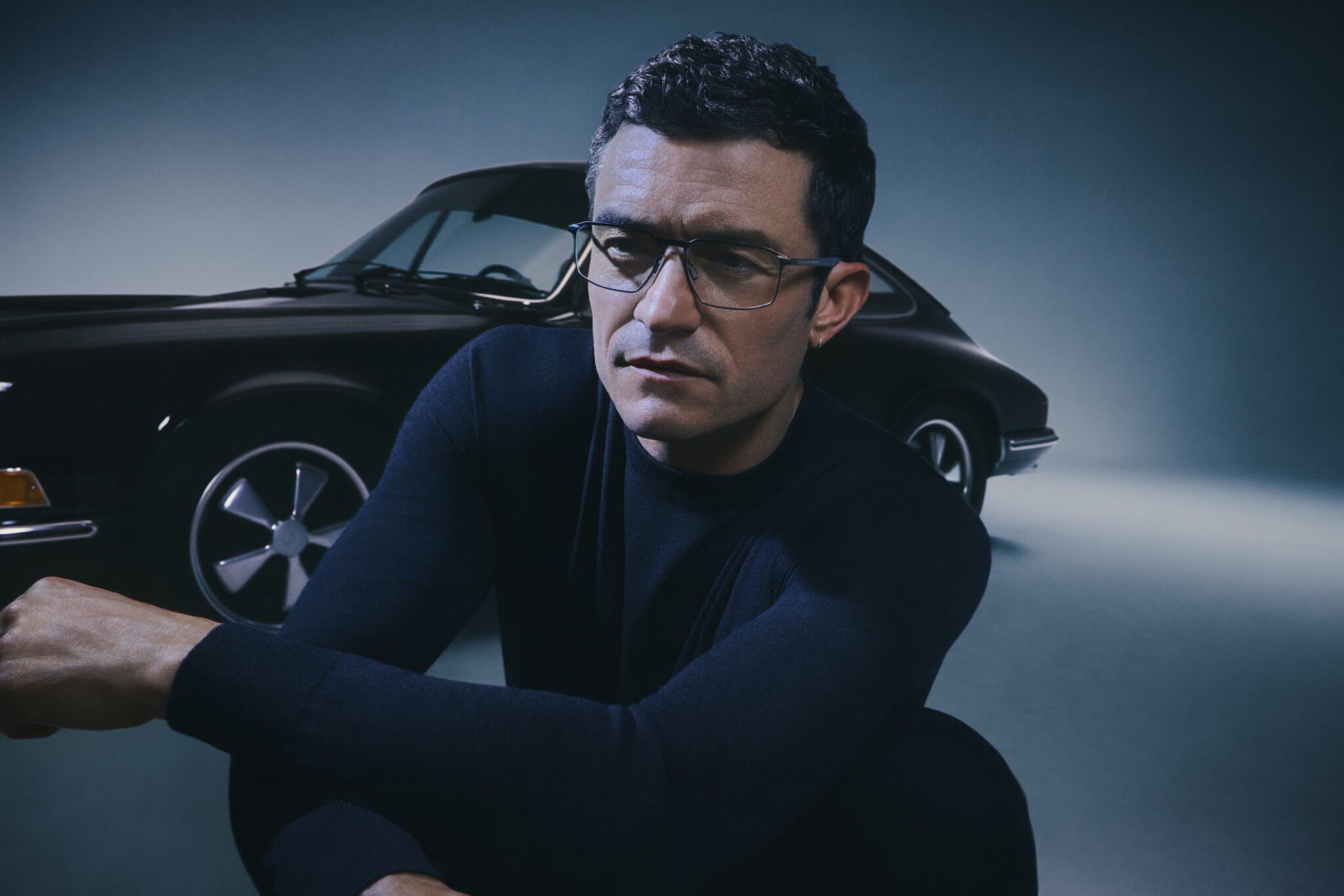 A person wearing glasses and a dark sweater sits in front of a vintage car with the focused demeanor of a Porsche Design brand ambassador.