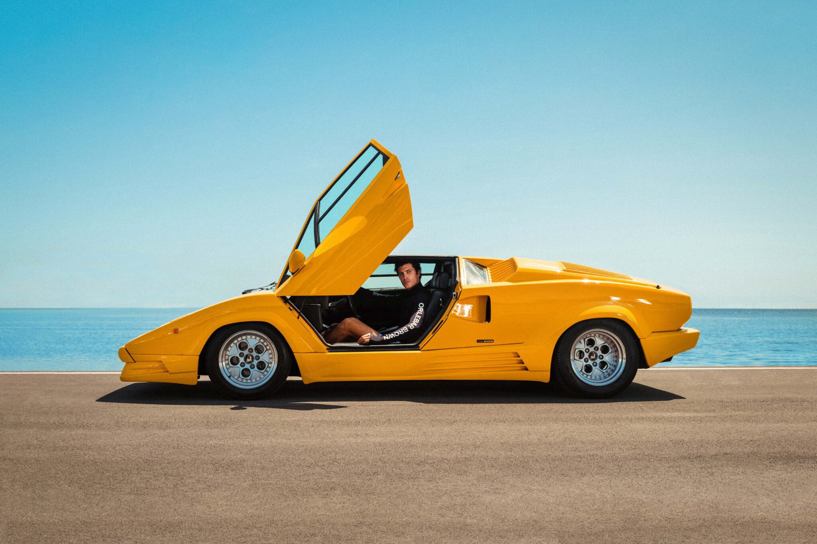 A man sits in a yellow Lamborghini with the doors open, exuding style reminiscent of the Miami Beach Collection.