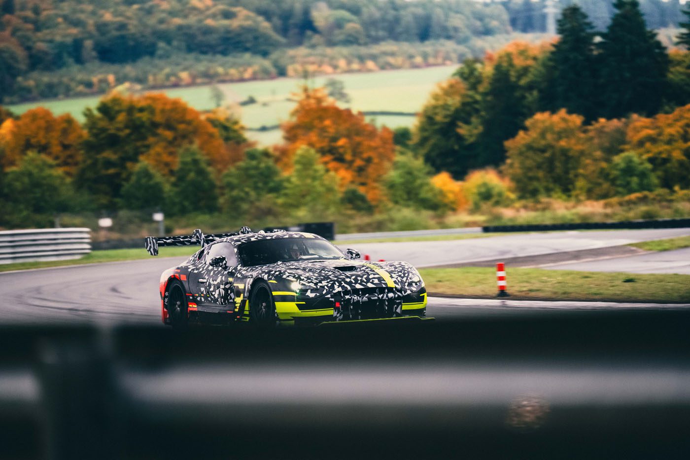 A camouflaged Mercedes CONCEPT AMG TRACK SPORT with yellow accents speeds along a racetrack, weaving through circuits bordered by trees in vibrant autumn foliage.