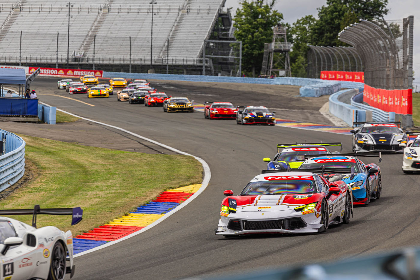 A group of race cars, including the Ferrari of Seattle, speed around a curved section of a racetrack, with empty grandstands and green trees in the background during the Ferrari Challenge.