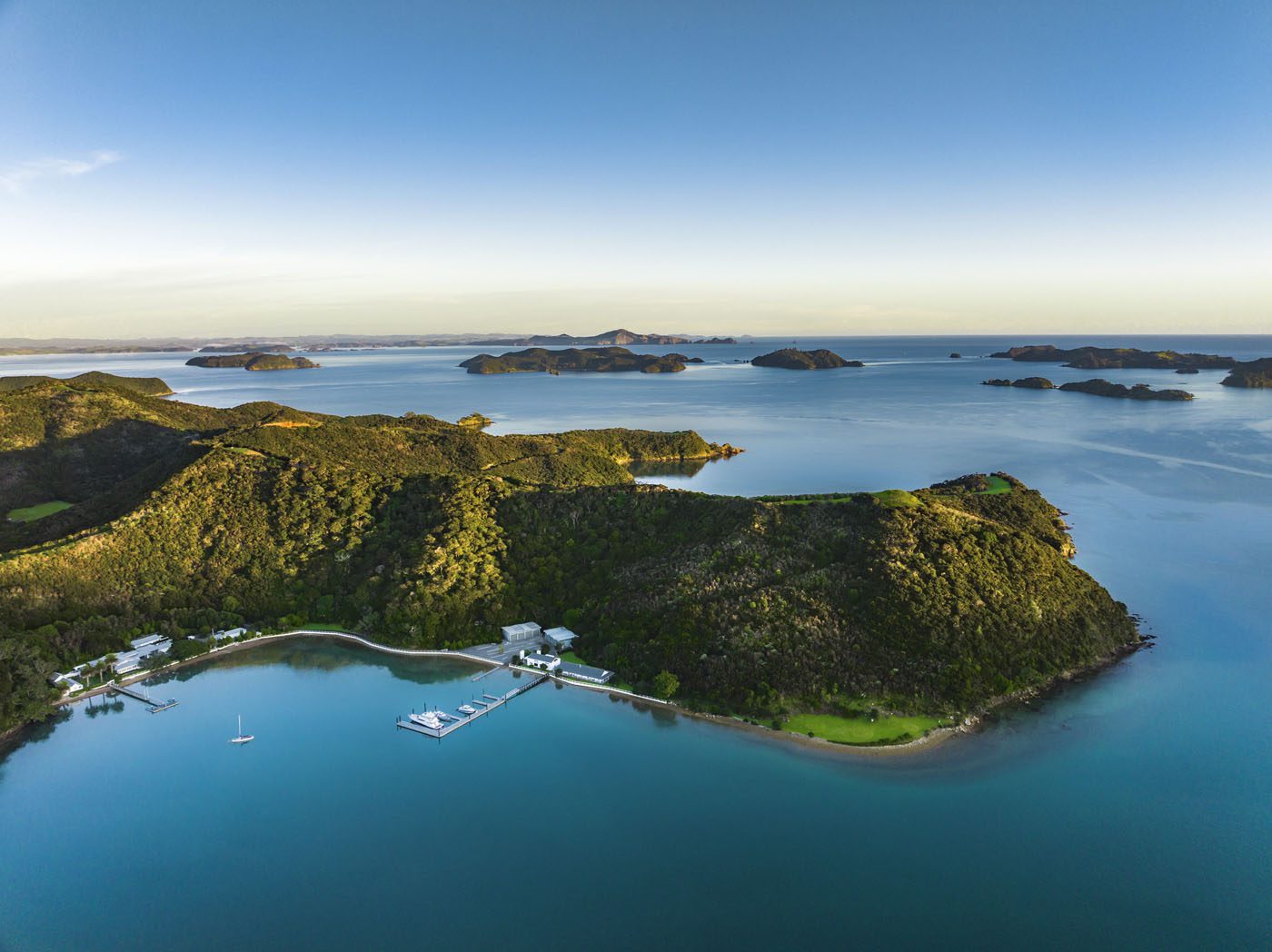 Aerial view of a lush, green peninsula along the coveted coastline, surrounded by calm blue water, with docks, buildings, and small islands visible in the distance under a clear sky.