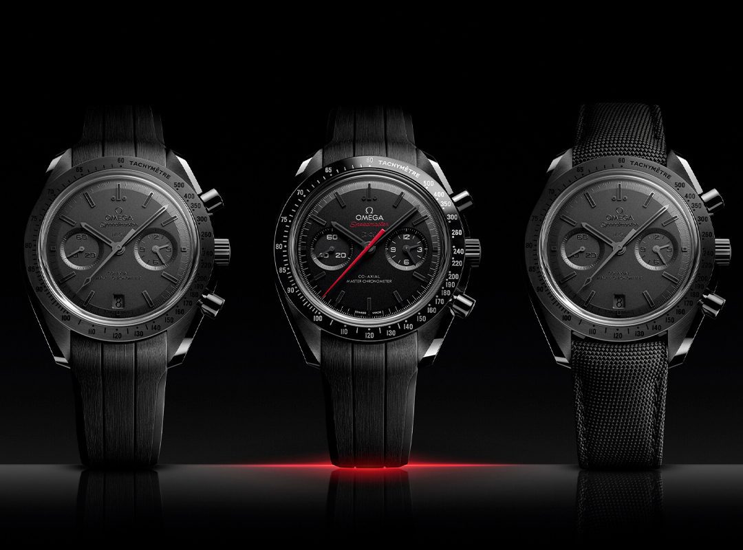 Three black Omega Speedmaster chronograph watches are displayed side by side against a dark background, with the center watch featuring a red second hand.