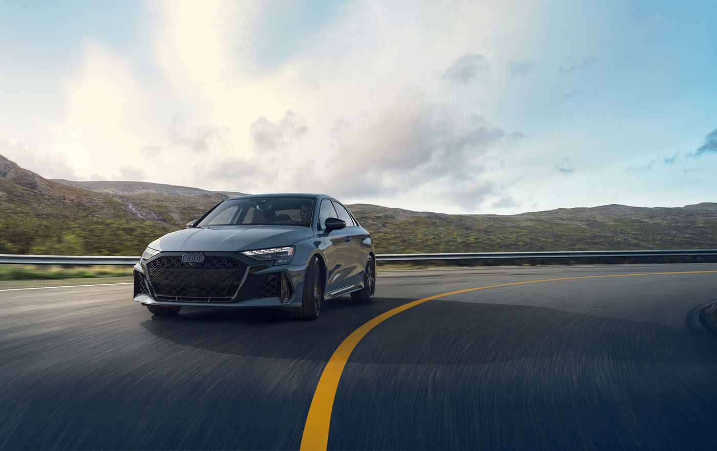 Under a cloudy sky, the sleek 2025 RS 3 confidently takes a sharp turn on a winding mountain road, showcasing Audi's impressive 5-cylinder legacy.