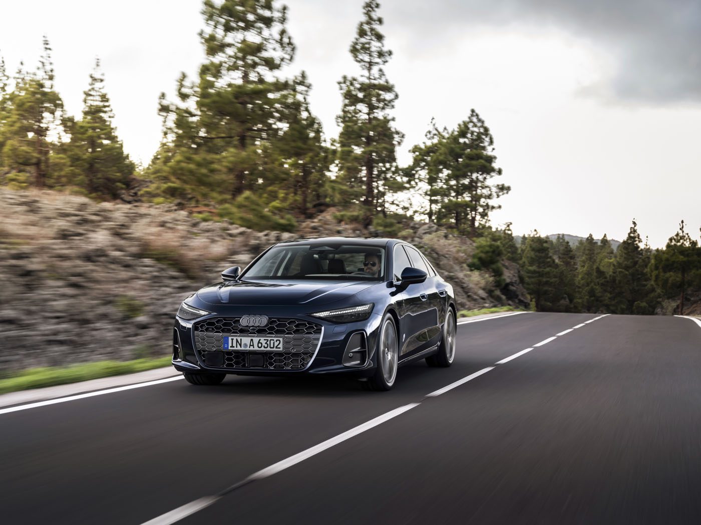 A dark blue new Audi A6 sedan glides along a winding, empty road bordered by trees and rocky terrain under a cloudy sky.