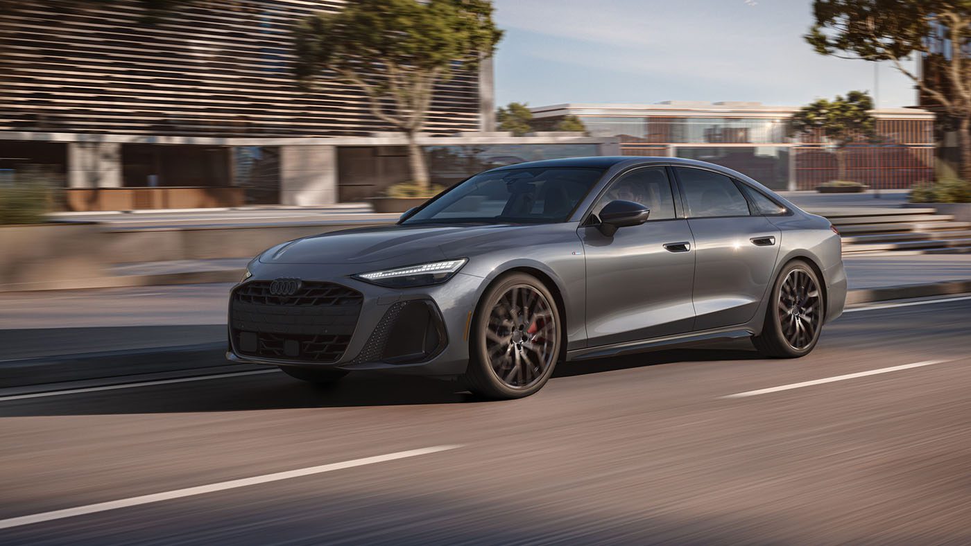A sleek gray 2026 Audi A6 with black wheels glides down a city street beside modern buildings and trees, under daylight.