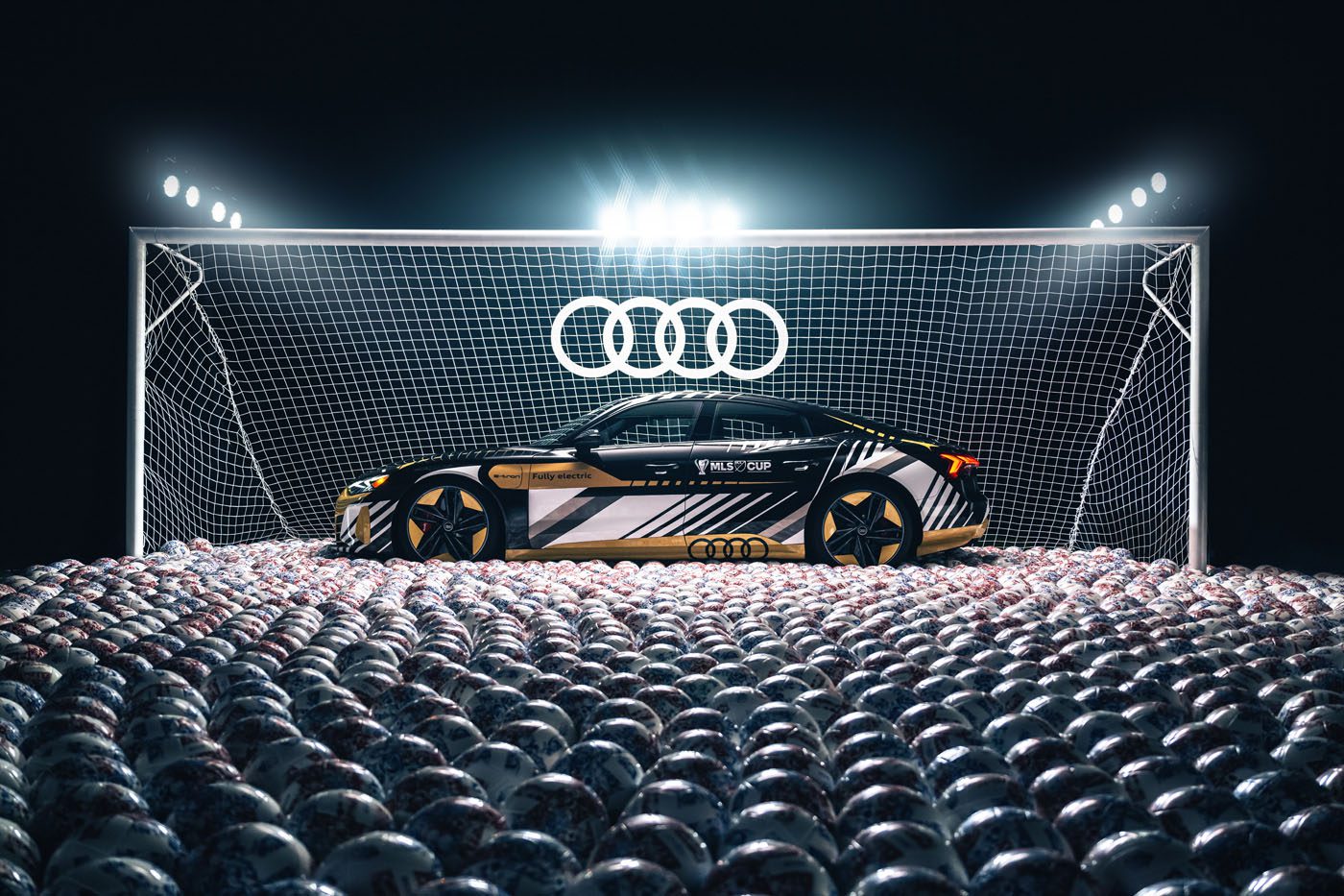 A black and gold Audi sports car is parked in front of a goalpost filled with soccer balls under bright stadium lights, celebrating the Audi partnership with Major League Soccer, with an illuminated Audi logo above the net.