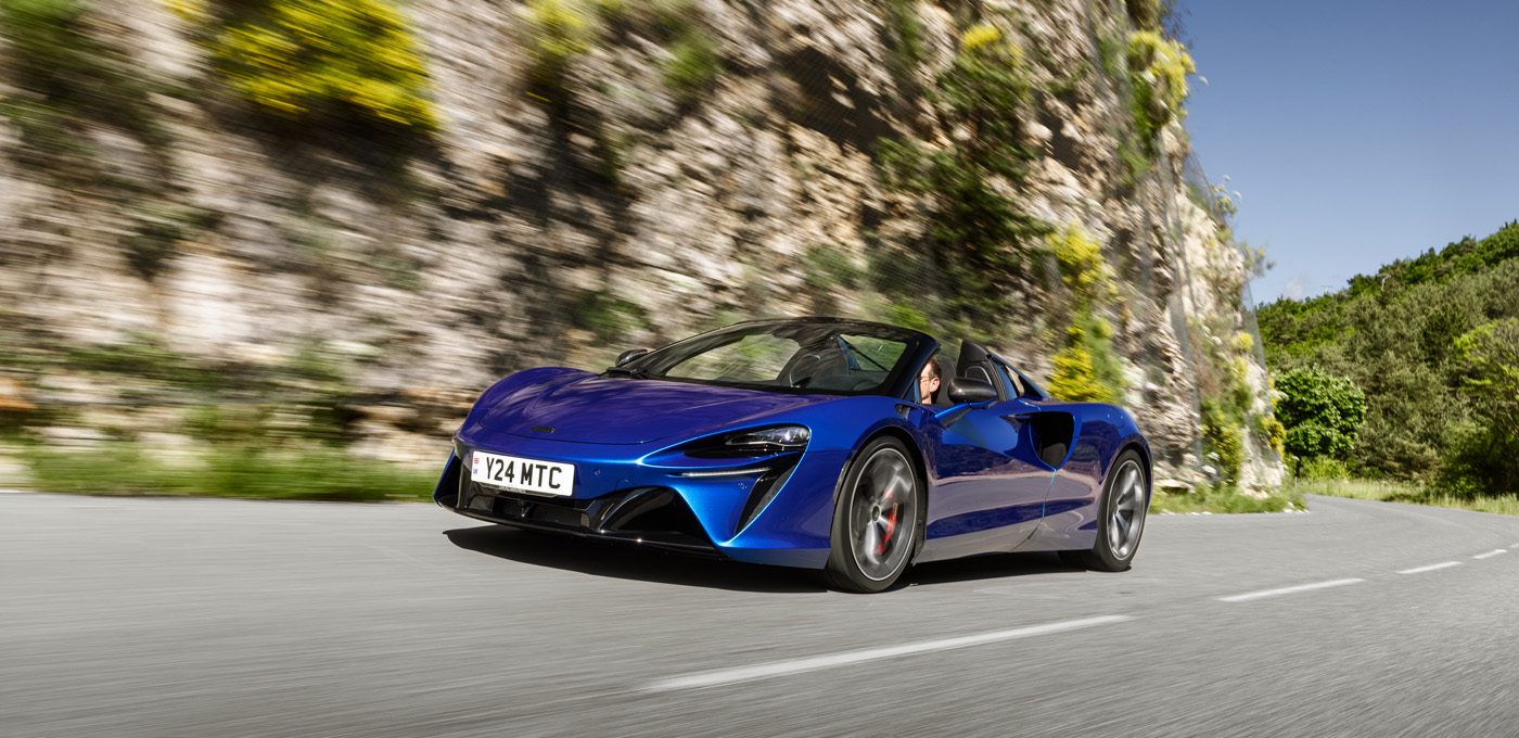 A blue sports car with a convertible top, reminiscent of a McLaren Trophy America series model, drives on a winding road next to rocky terrain and greenery under a clear blue sky.