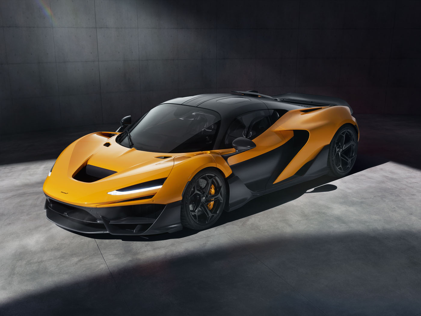 A sleek, orange and black McLaren W1 sports car with a 3D printed suspension system is parked on a concrete floor in a dimly lit room.